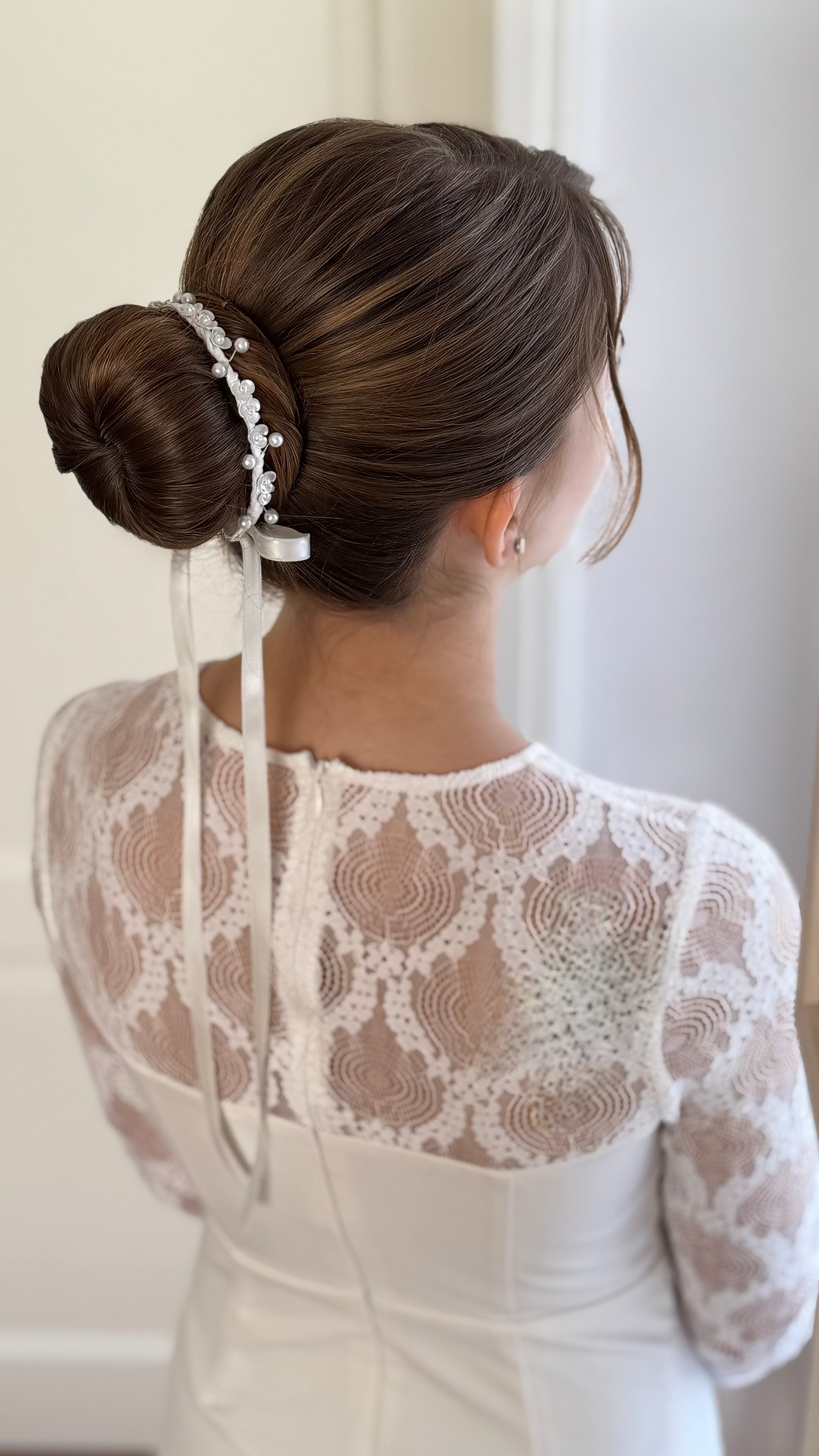 Mädchen im weißen Kleid mit festlicher Duttfrisur, dekoriert mit Blumen-Perlen-Haarschmuck für die Erstkommunion.