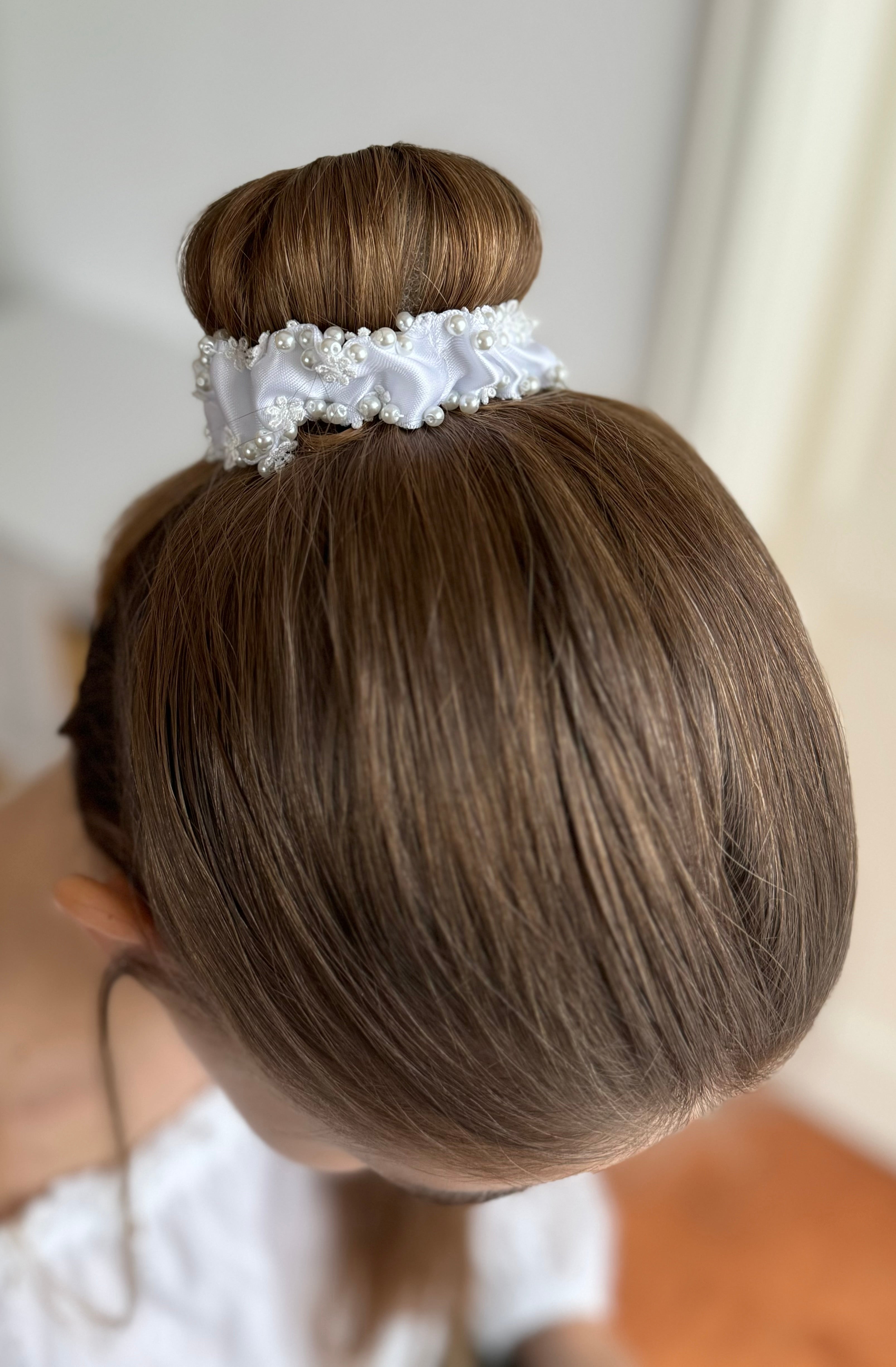 Ein Dutt, dekoriert mit einem weißen handgefertigten Haarschmuck aus Stoff, Spitze und Perlen. Der Haarschmuck liegt eng und gleichmäßig um den Dutt.